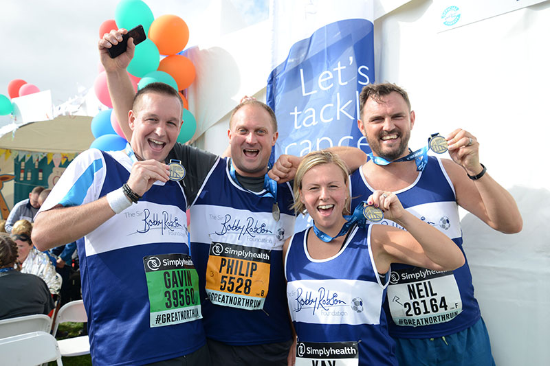 4 runners holding up medals celebrate in the Sir Bobby Foundation tent at the end of the Great North Run